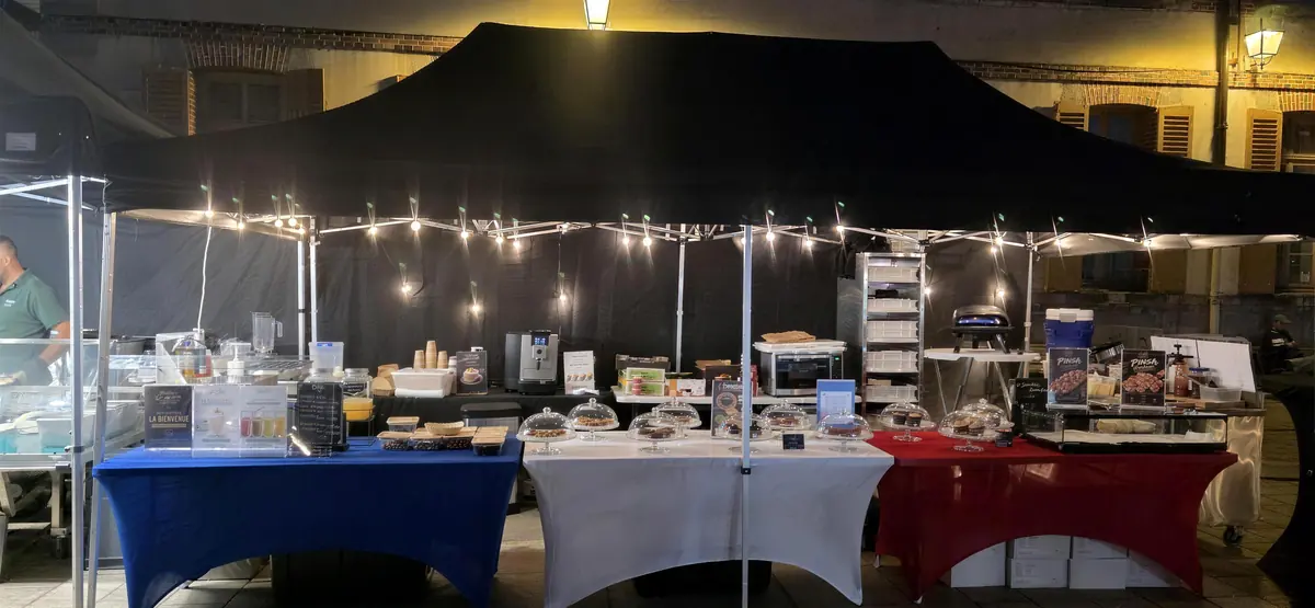 Le buffet du stand, sous un barnum noir éclairé de guirlandes, avec des gourmandises sous cloches de verre